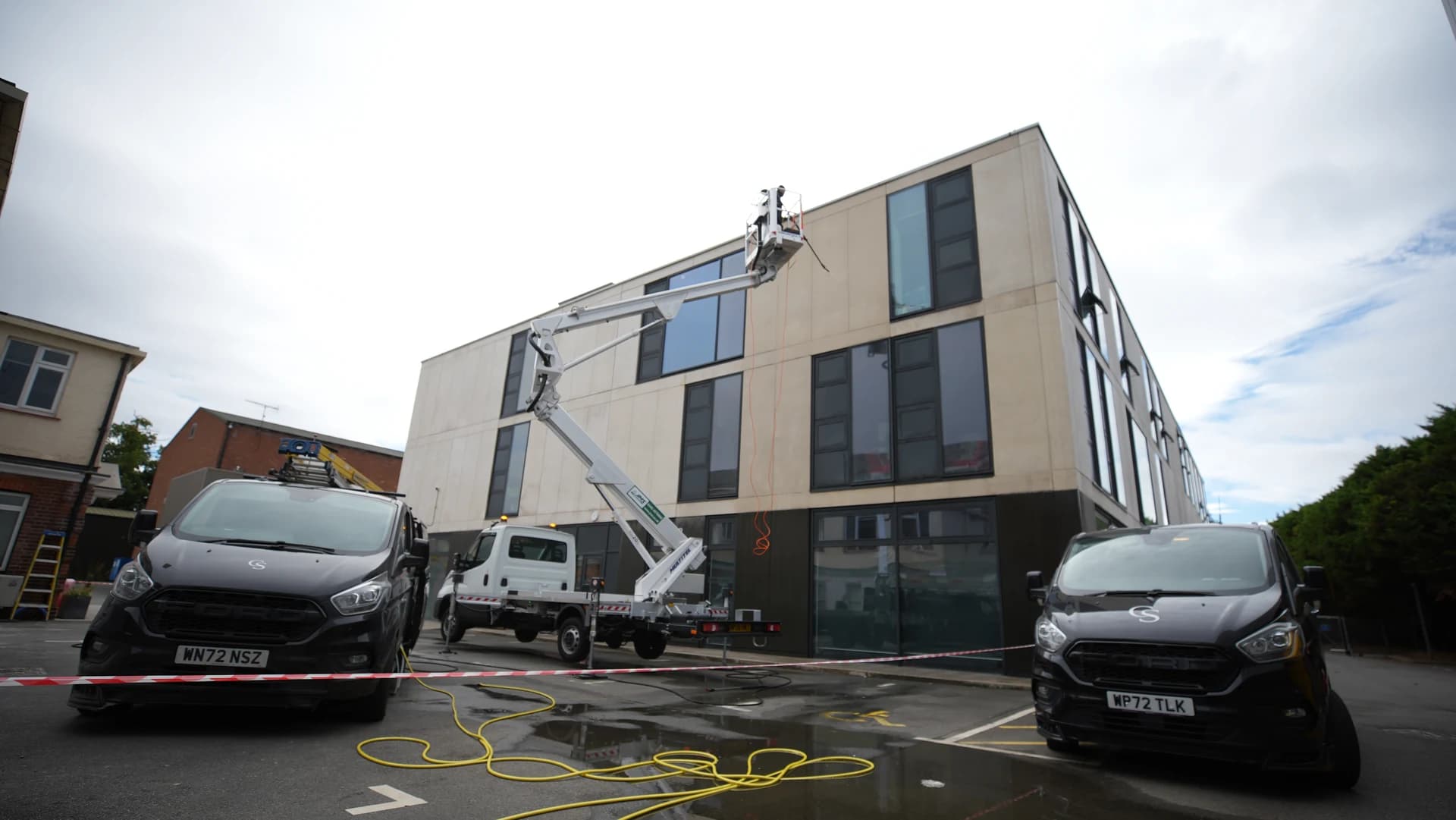 Wide view across barrier tape to a truck-mounted access platform reaching the top of a building elevation, a van on each side