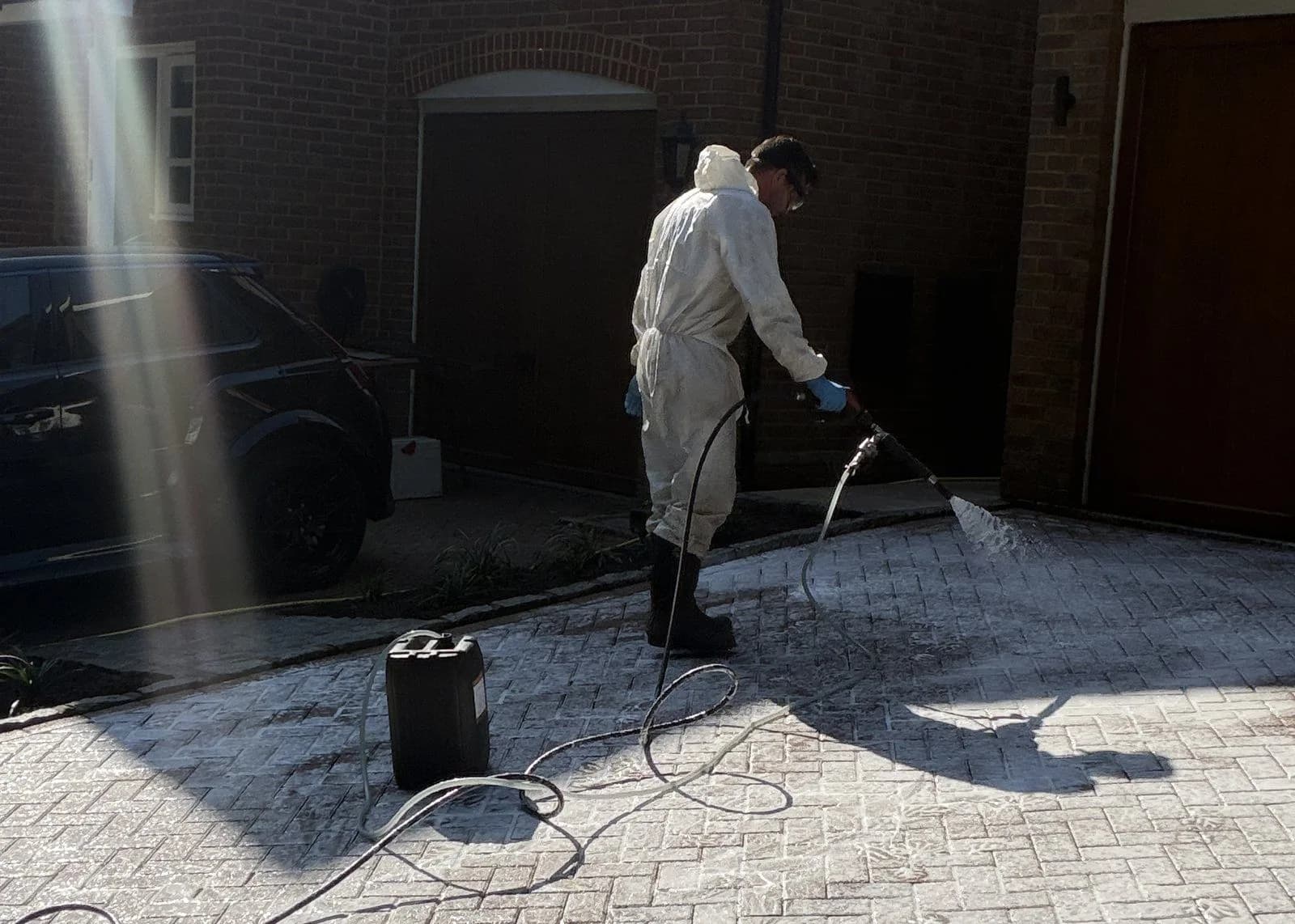 Chiltern Surface Co technician in protective coveralls spraying the chemical treatment across the block paved driveway in Hazlemere