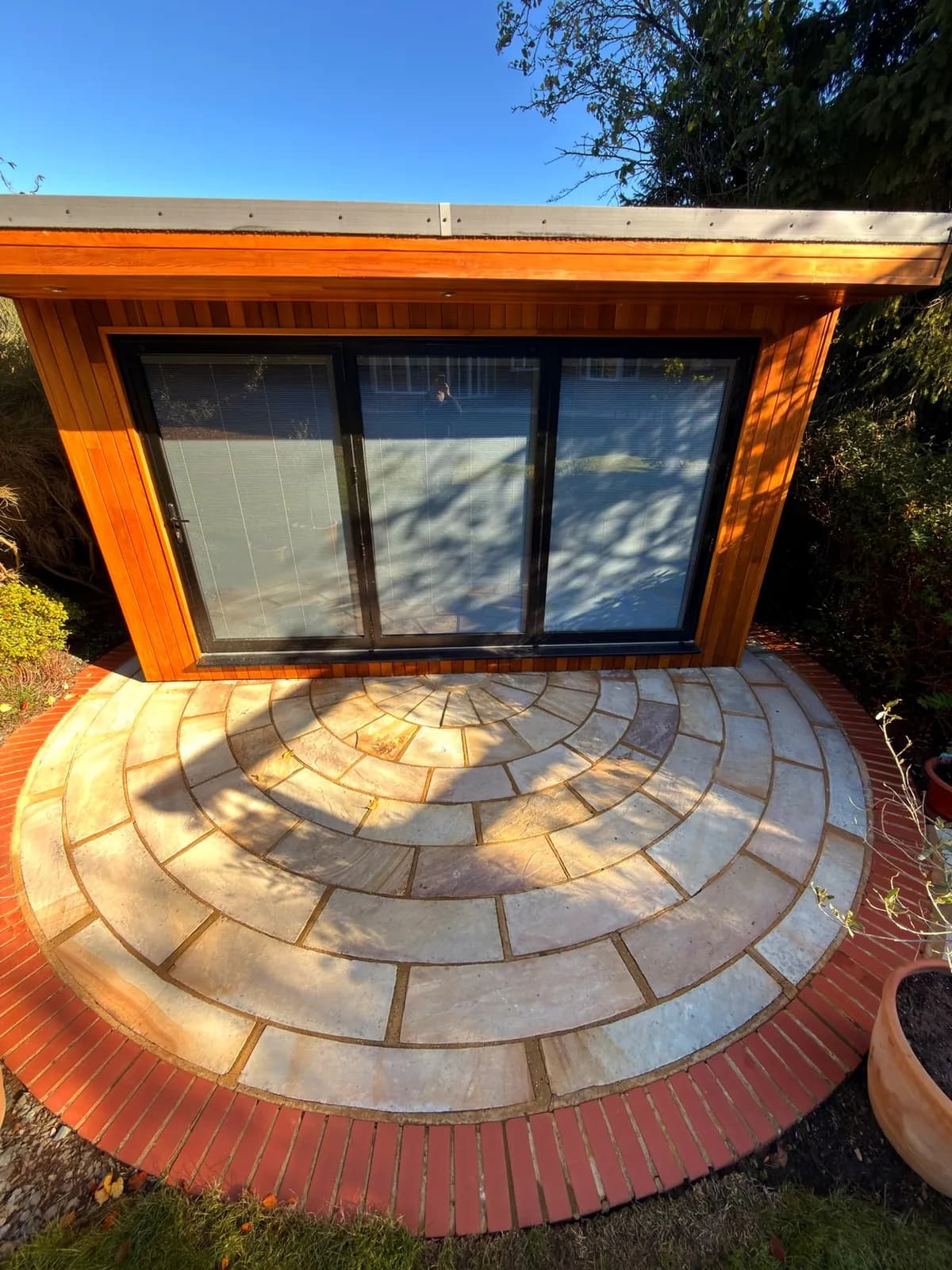The circular Indian sandstone terrace in front of the cedar garden room after cleaning, framed by red brick edging under a blue sky