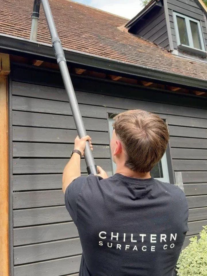 Operative in a branded T-shirt raising a carbon gutter vacuum pole to the eaves of a clay-tiled, weatherboarded building