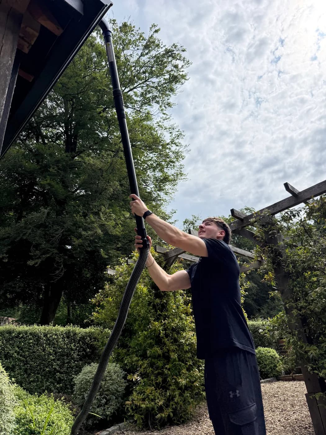 Low view of an operative raising a black gutter vacuum pole and hose to the eaves of a timber building beneath tall trees