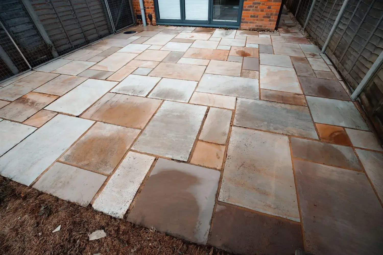 Damp sandstone patio drying unevenly in a fenced rear garden, outside grey patio doors
