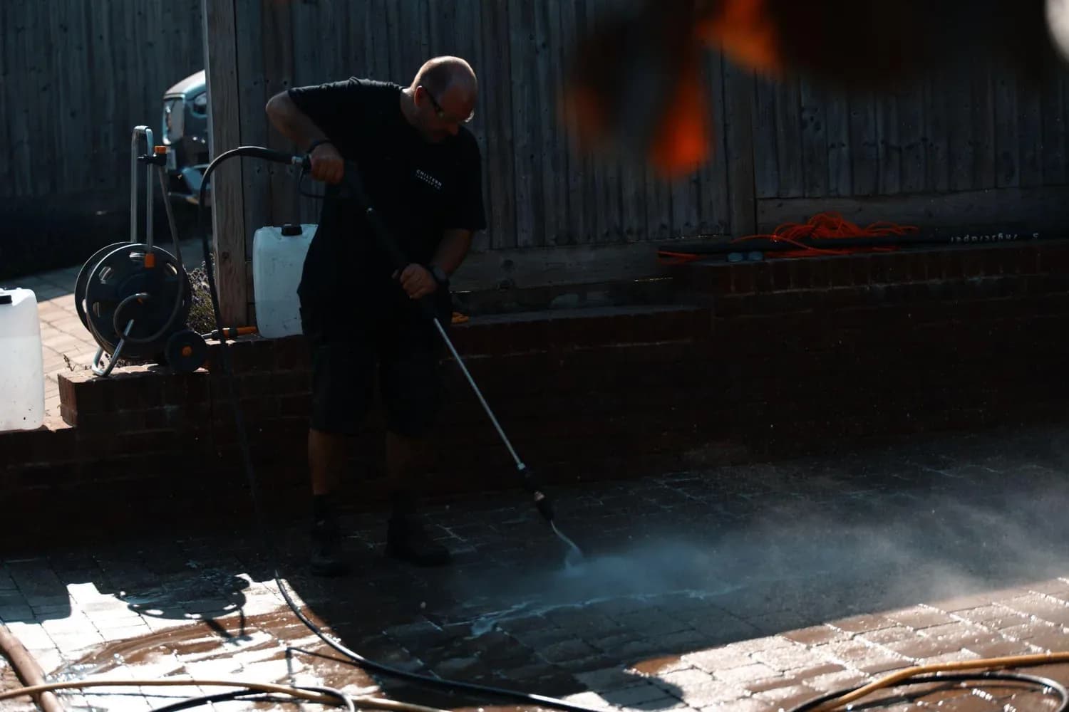 Operative in a Chiltern Surface Co T-shirt pressure washing dark block paving, with hose reel and containers behind