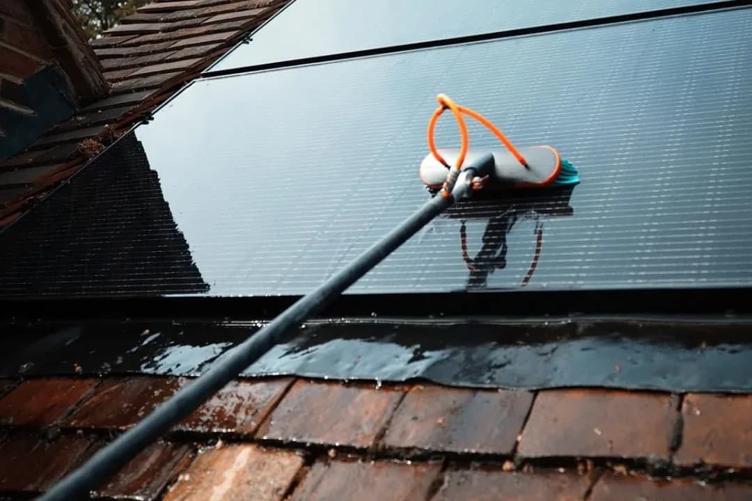 Water-fed brush cleaning wet black solar panels fitted to a clay-tiled roof below a roof window