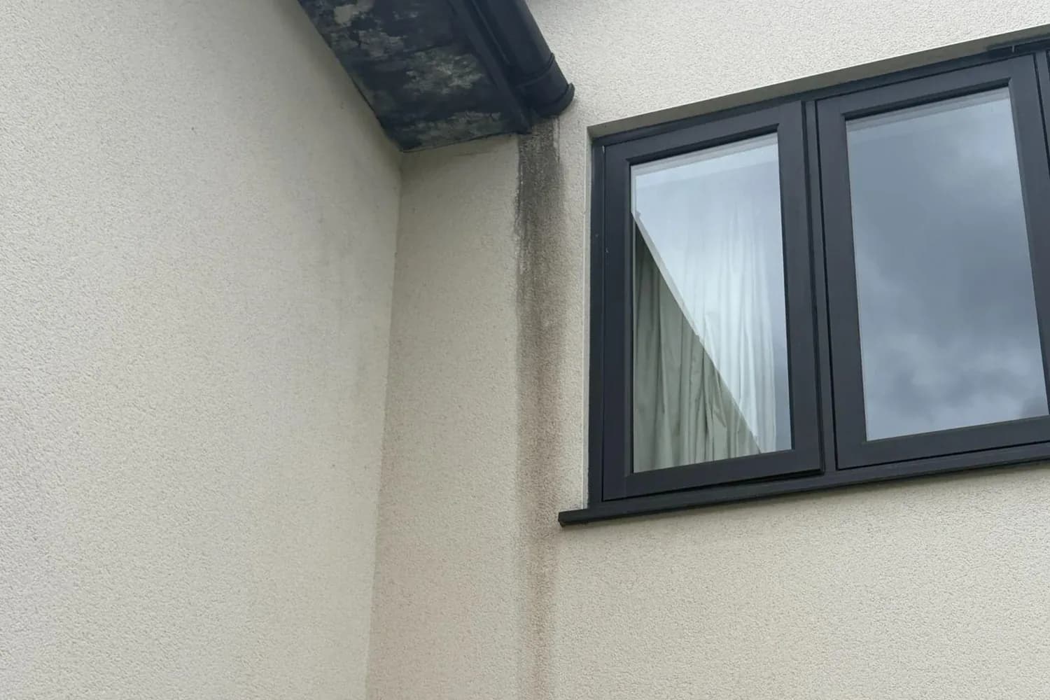 Cream textured render beneath a black-stained soffit, with a dark green-grey streak running down from the gutter end